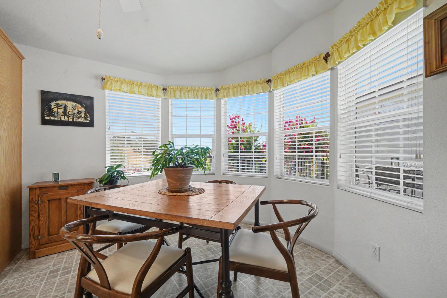 Dining room with a wooden table and chairs beside bright bay windows overlooking flowering shrubs.