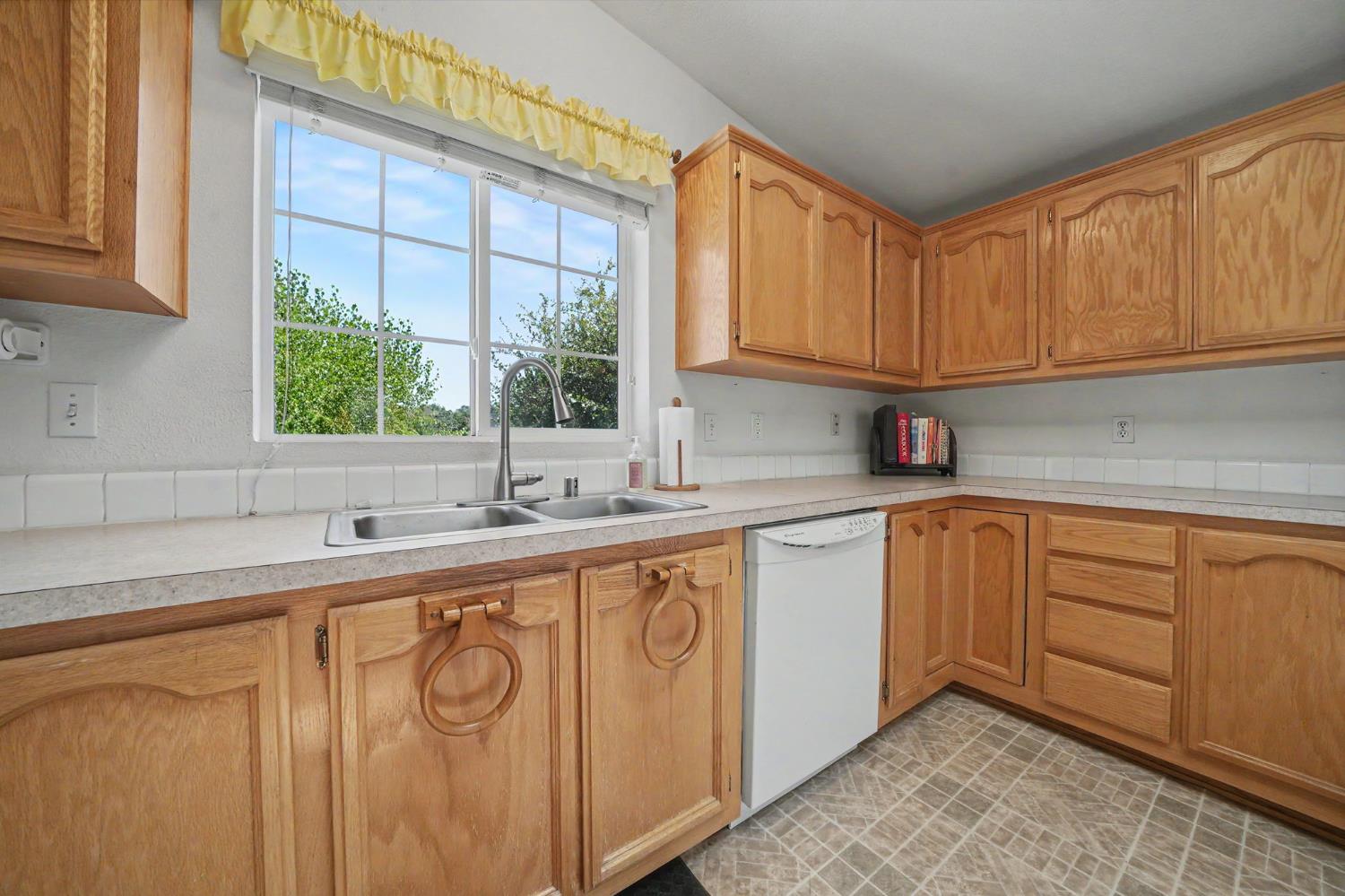 Kitchen with honey-colored wood cabinets, a double sink beneath a large window, and a white dishwasher.