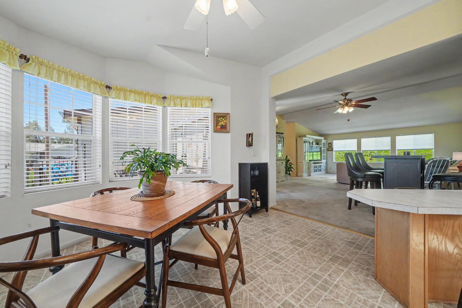 Dining area with a wooden table beside wide windows, opening into a carpeted living room.