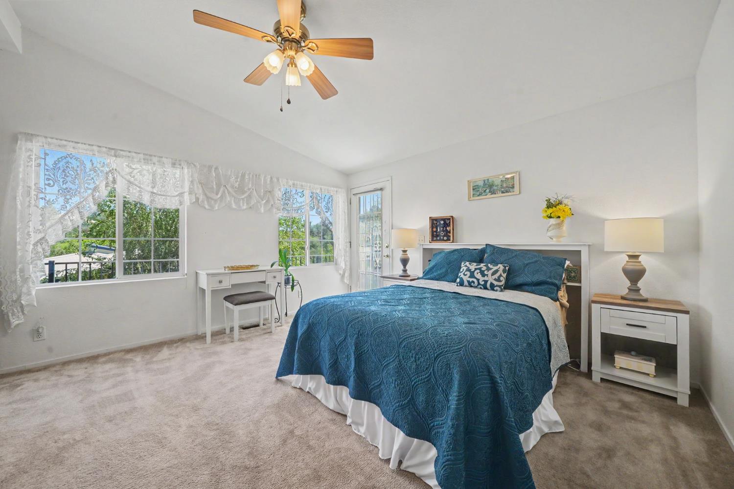 Staged bedroom with a blue quilted bed, lace-curtained windows, and a wood-blade ceiling fan.