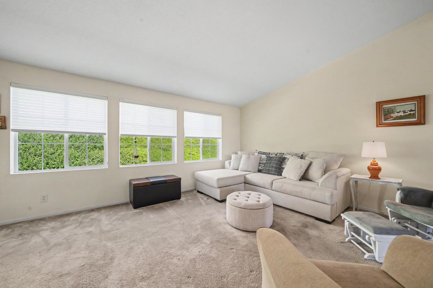 Living room with a cream sectional sofa, round ottoman, and three windows overlooking greenery.