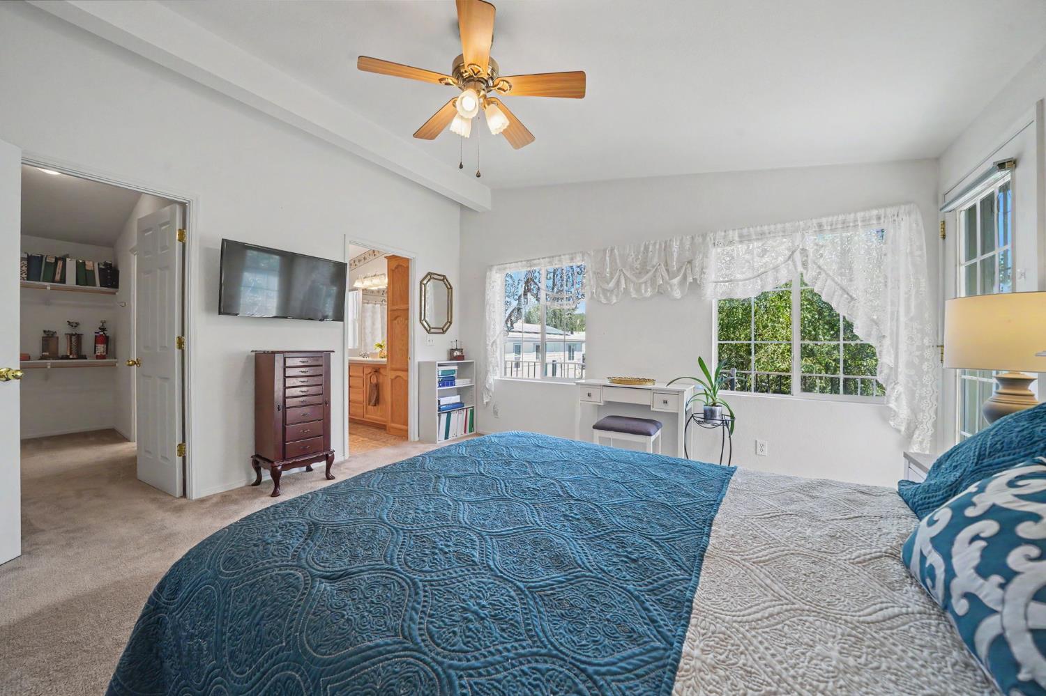Bedroom with a blue quilted bed, ceiling fan, and lace-curtained windows.