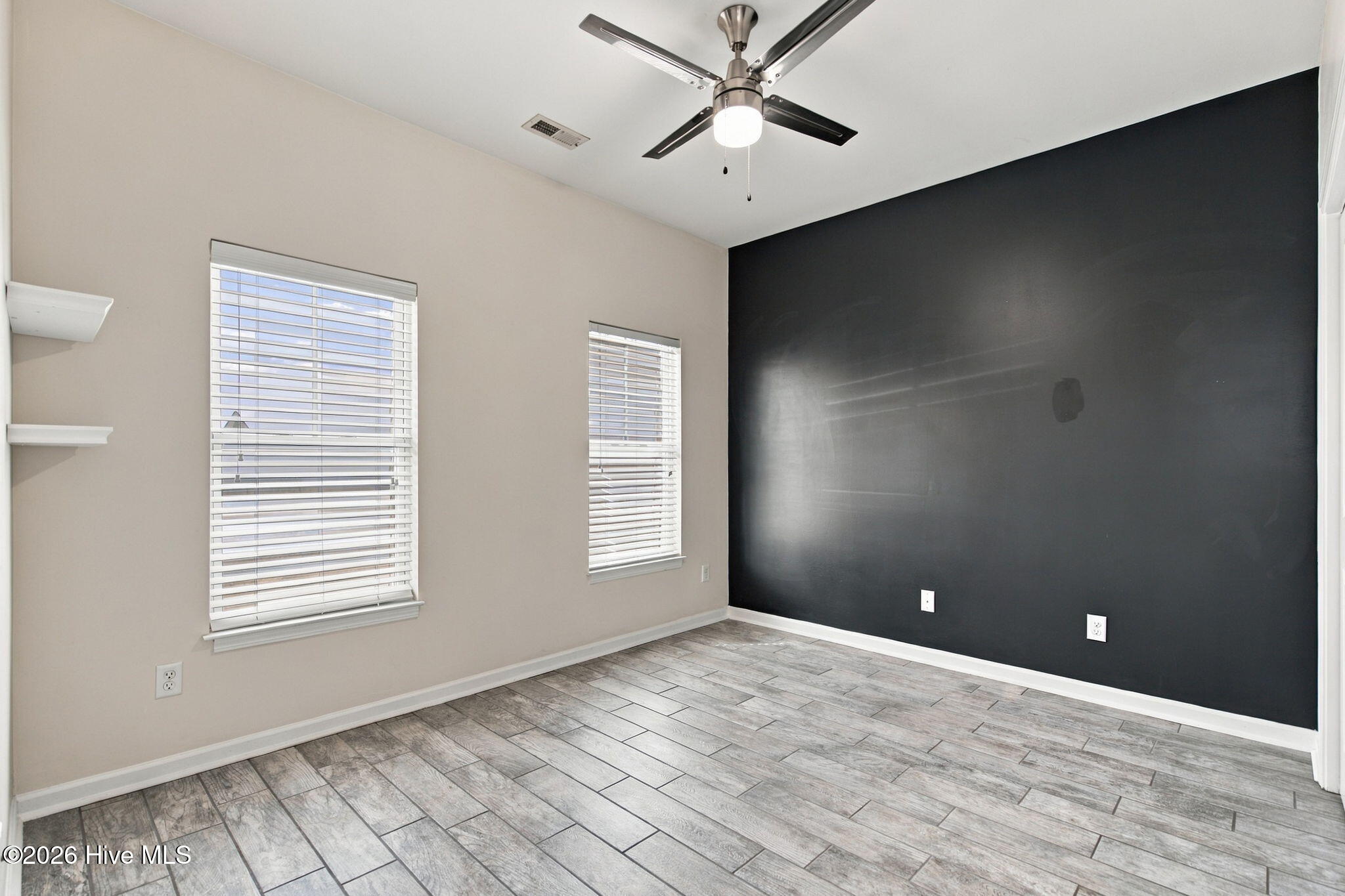 Empty room with gray tile flooring, a charcoal accent wall, two blinded windows, and a ceiling fan.