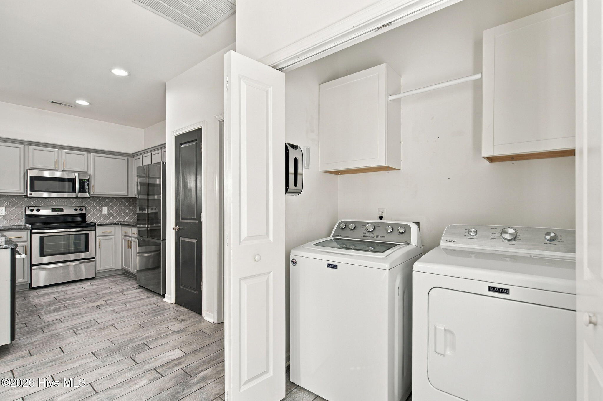 Laundry room with white washer and dryer, open cabinets, and an adjacent gray-and-stainless kitchen.