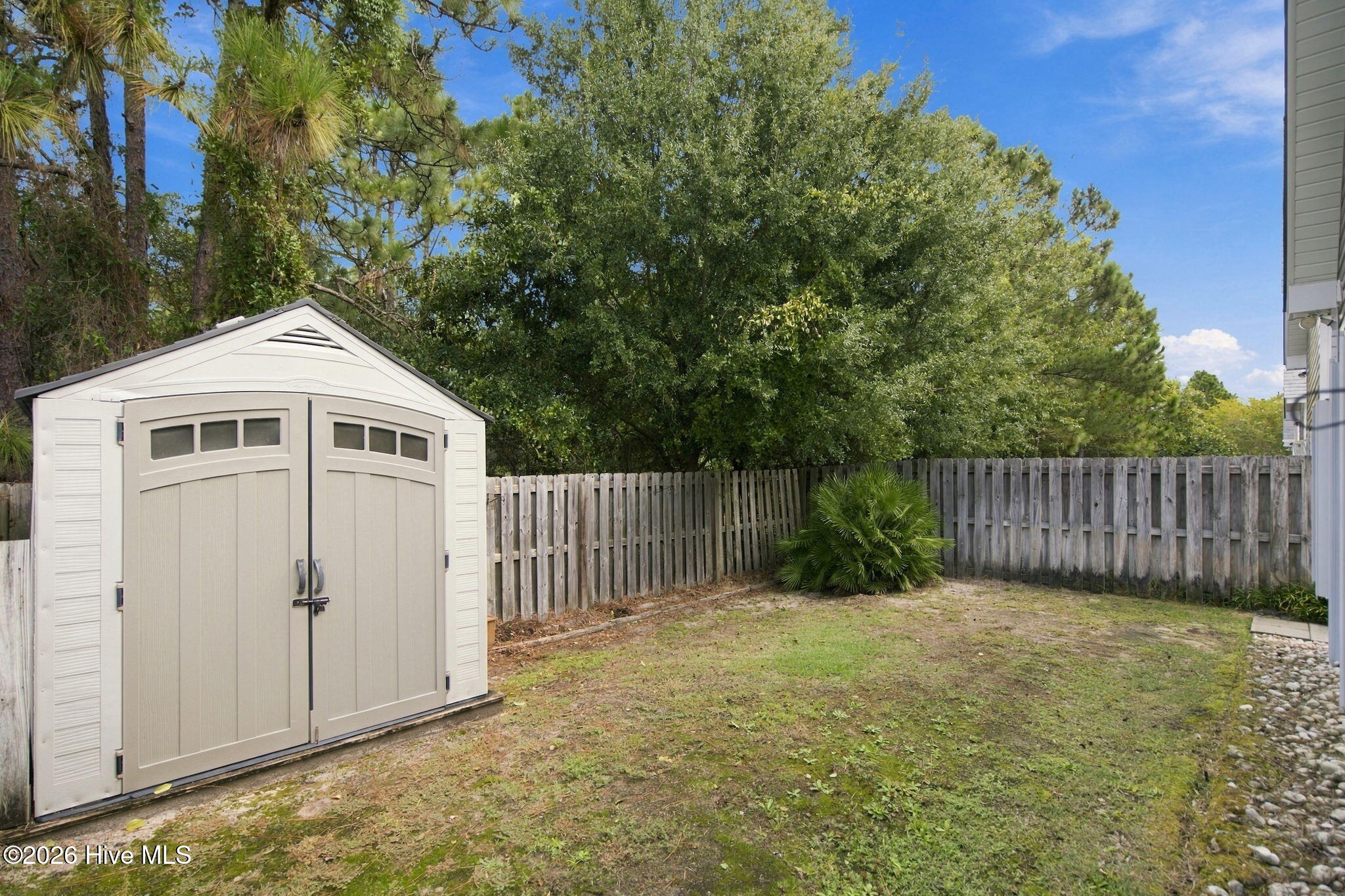 Fenced backyard with a beige storage shed, patchy lawn, and mature trees.