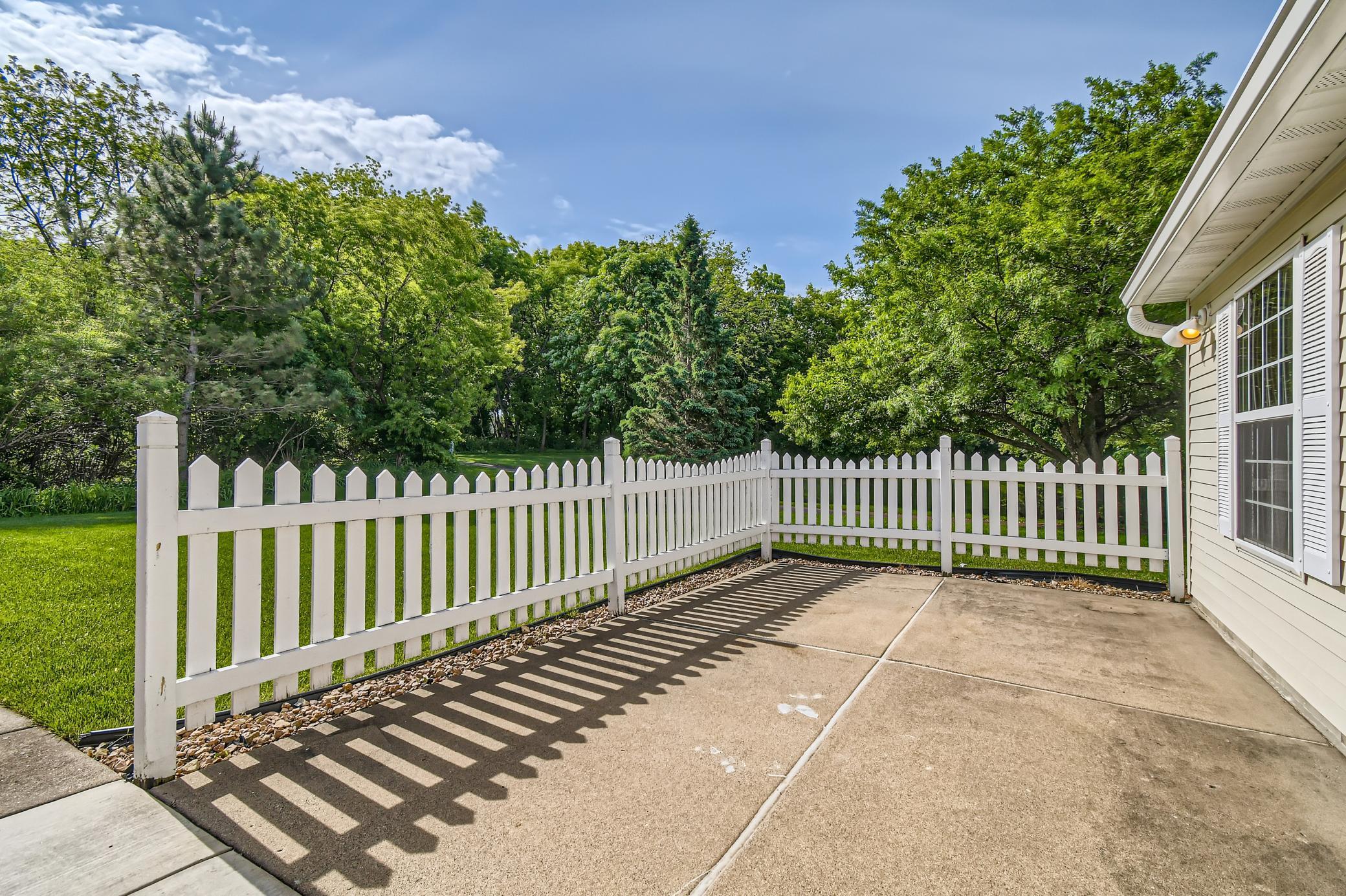 Concrete patio enclosed by a white picket fence, with tree-lined lawn beyond.