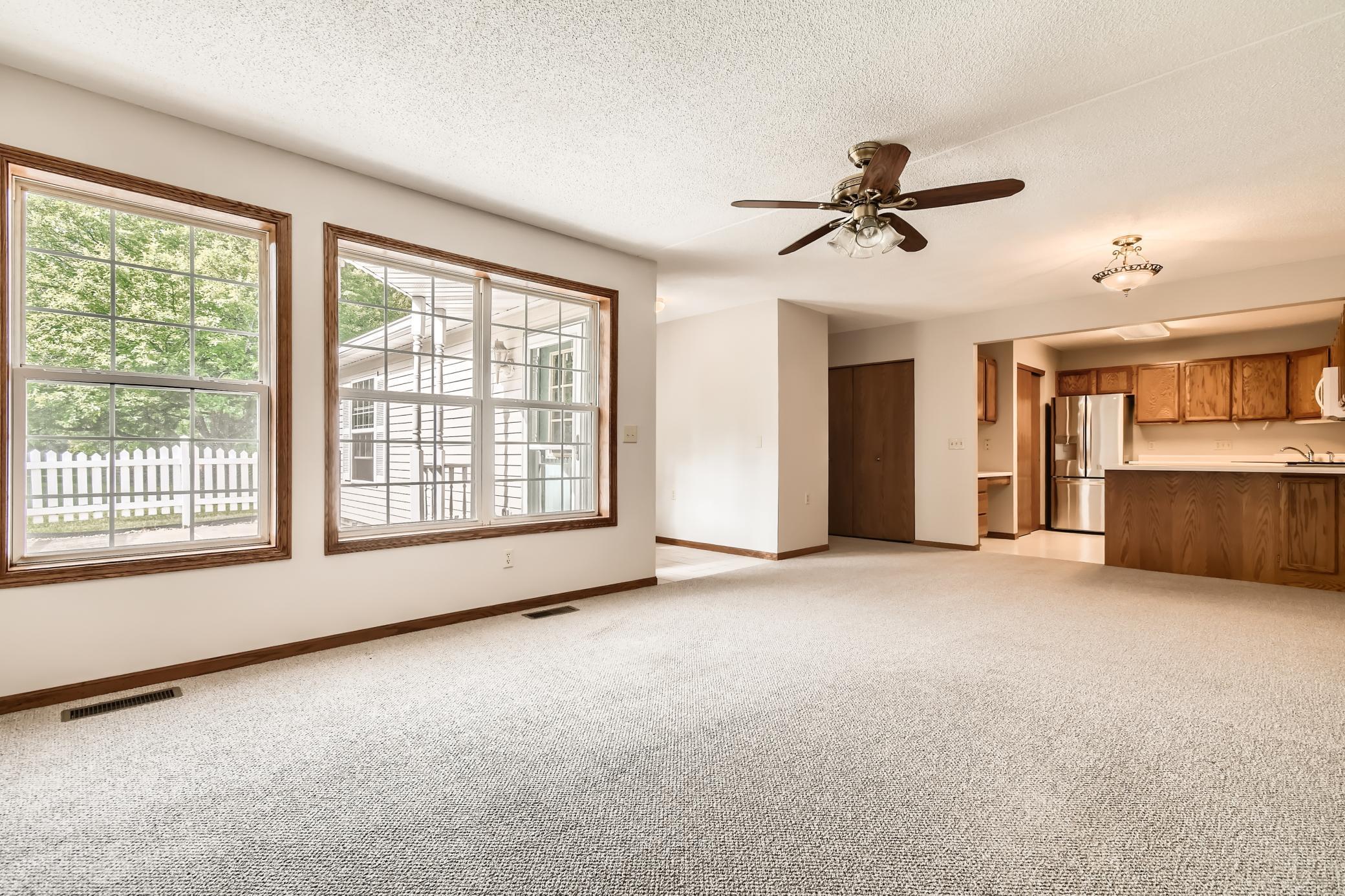 Empty carpeted living room opening to a kitchen, with large windows and a ceiling fan.