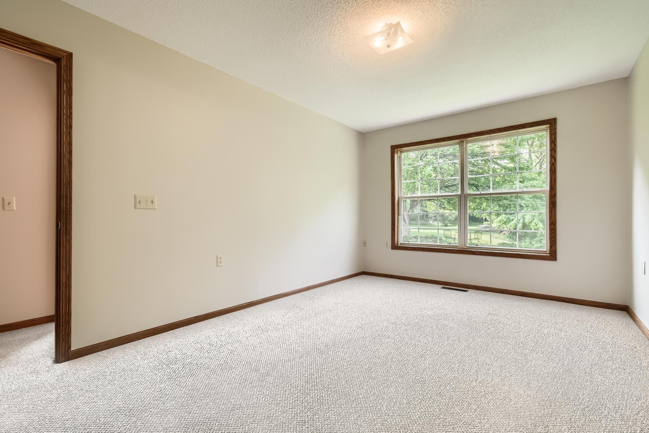 Empty carpeted room with dark wood trim and a large window overlooking trees and lawn.
