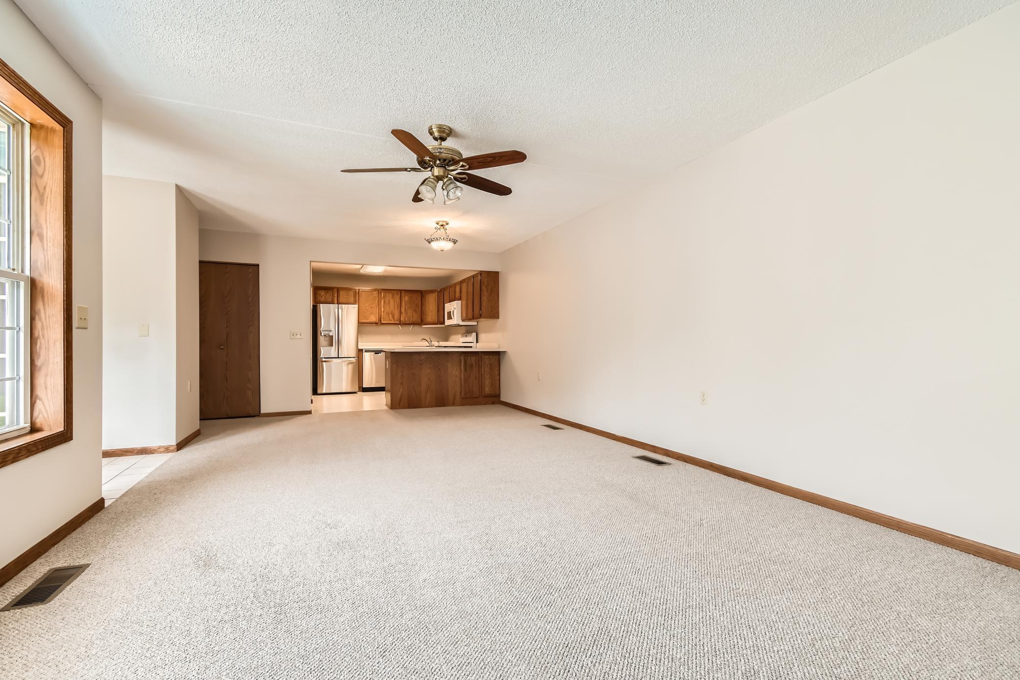Vacant carpeted living area with a ceiling fan and an open kitchen with wood cabinets.