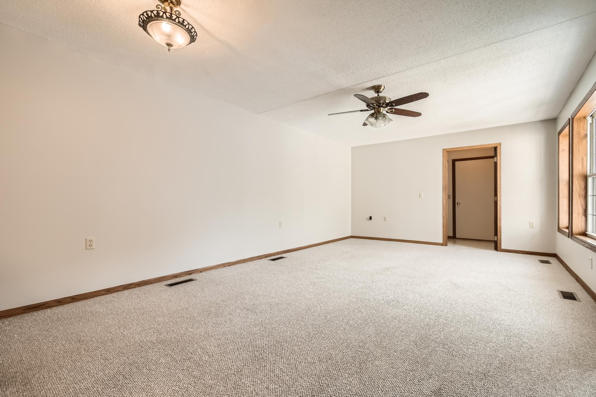 Empty carpeted room with a ceiling fan, wood trim, and windows along one wall.