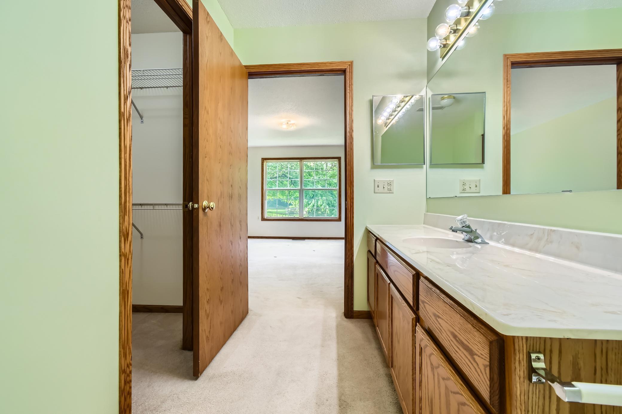 Bathroom with a long wood vanity and mirror, opening to a closet and a carpeted room.