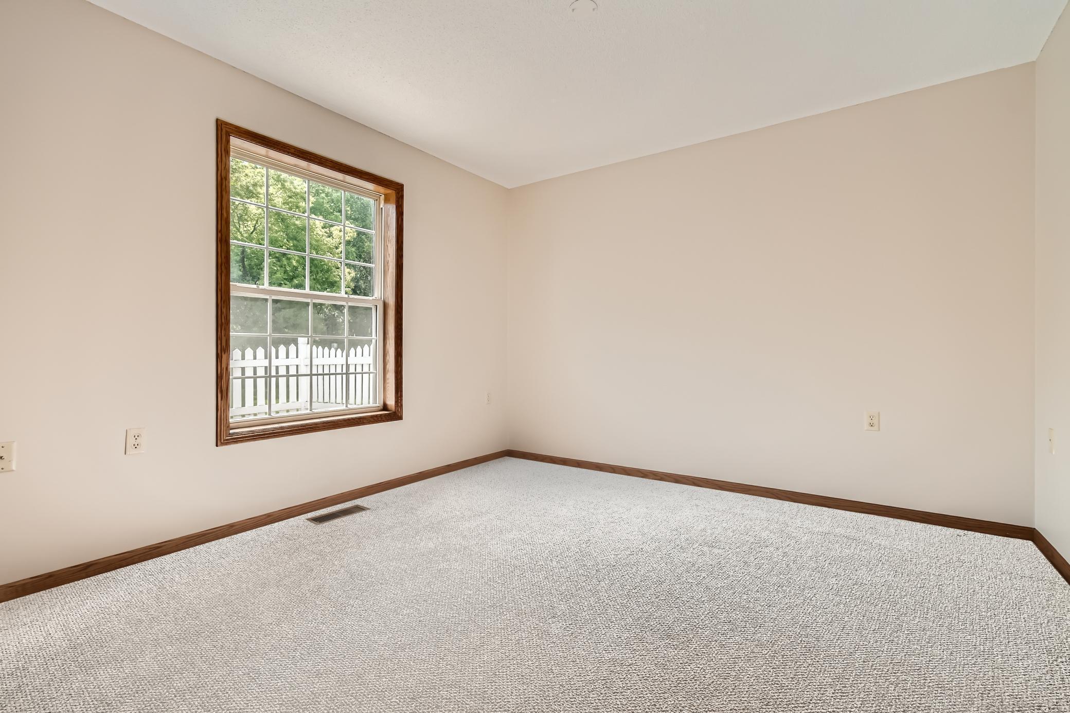 Empty bedroom with light carpet, dark wood trim, and a large window overlooking trees and a white fence.