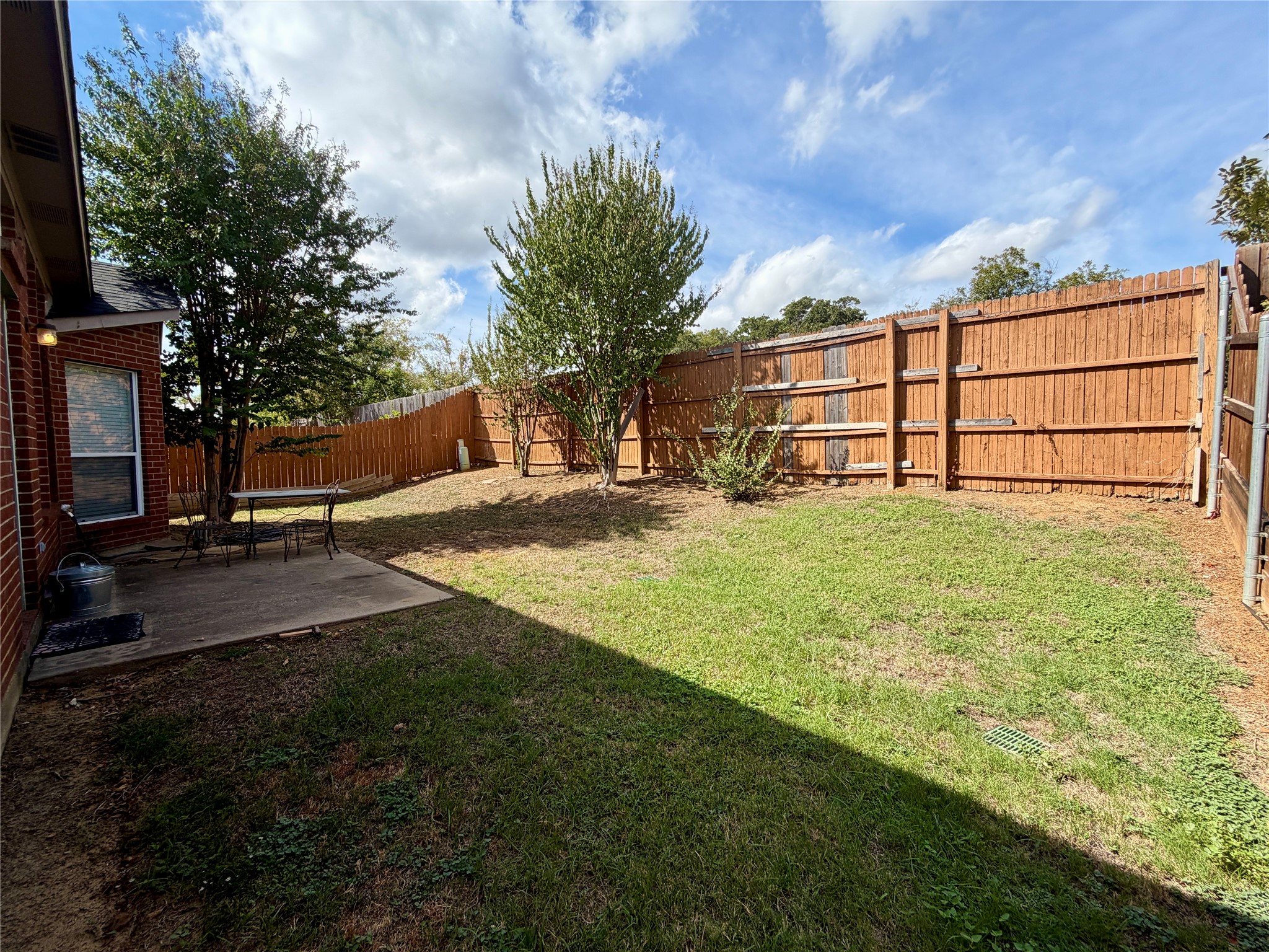 Fenced backyard with mature trees, a grassy lawn, and a concrete patio beside a brick house.