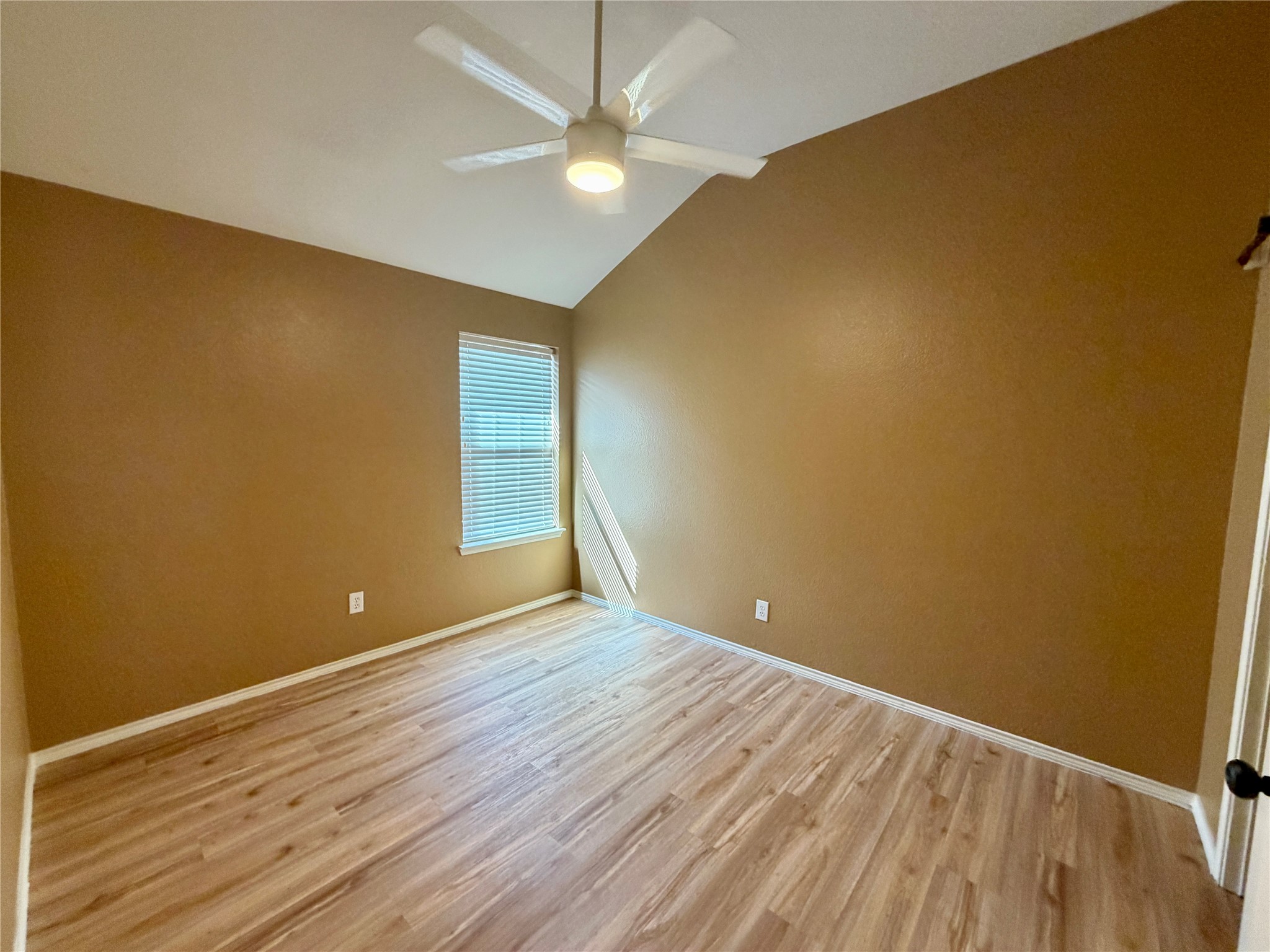 Empty bedroom with brown walls, wood-look flooring, and a ceiling fan; sunlight falls through closed blinds.