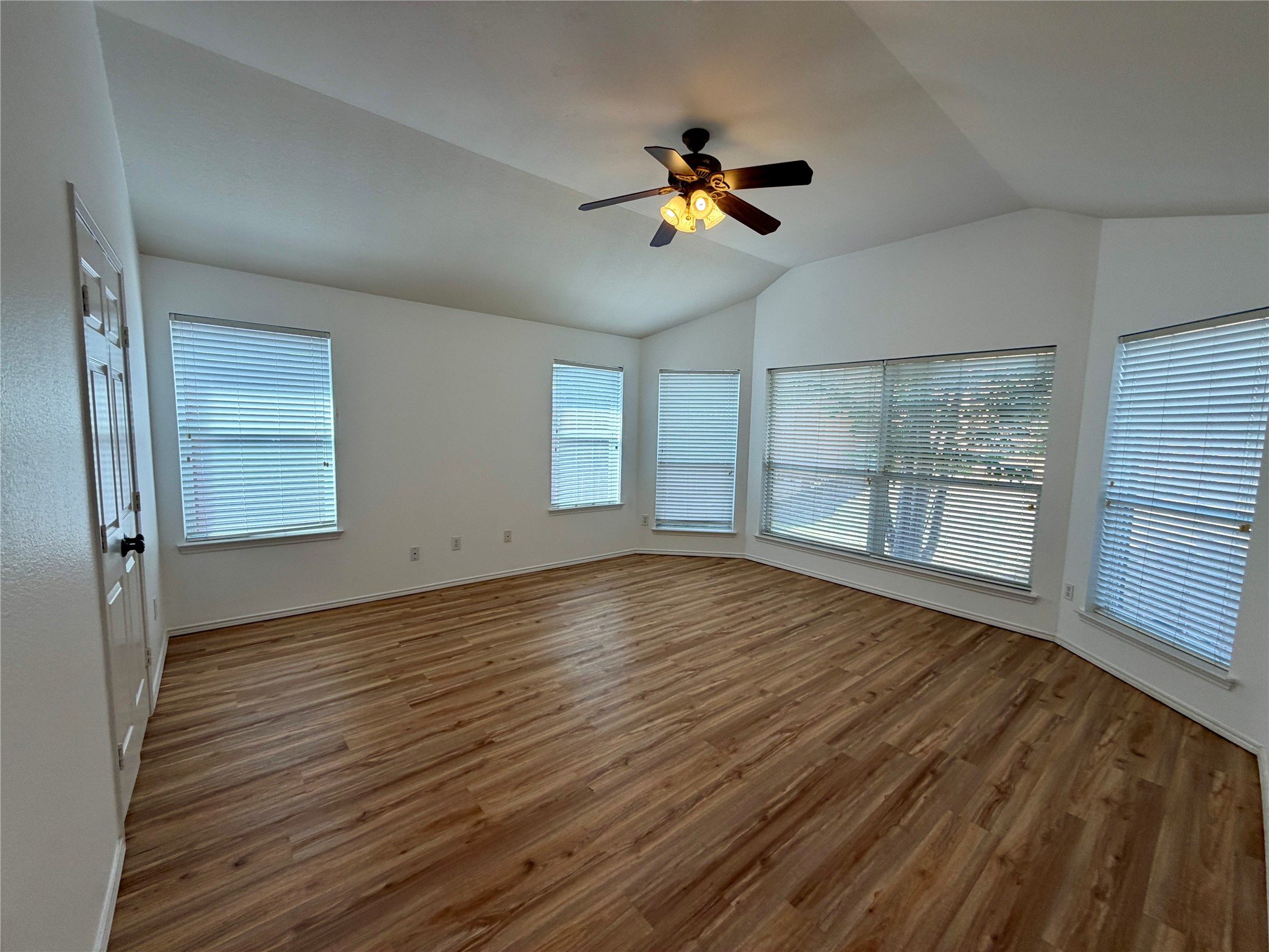 Vacant room with wood-look flooring, a vaulted ceiling, and multiple windows with blinds.