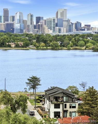 Multi-story home beside a lake, with a tree-lined shore and city skyline in the distance.