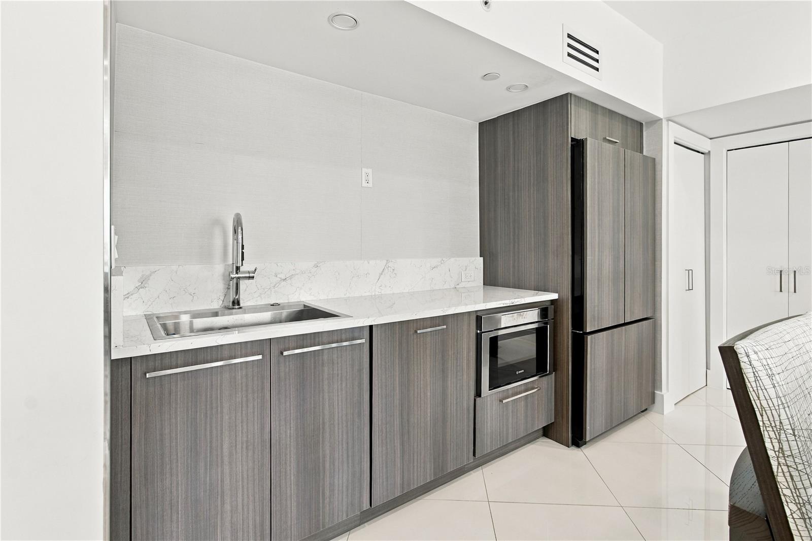 Modern condo kitchenette with dark cabinets, marble-look counters, stainless sink, built-in microwave, and refrigerator.