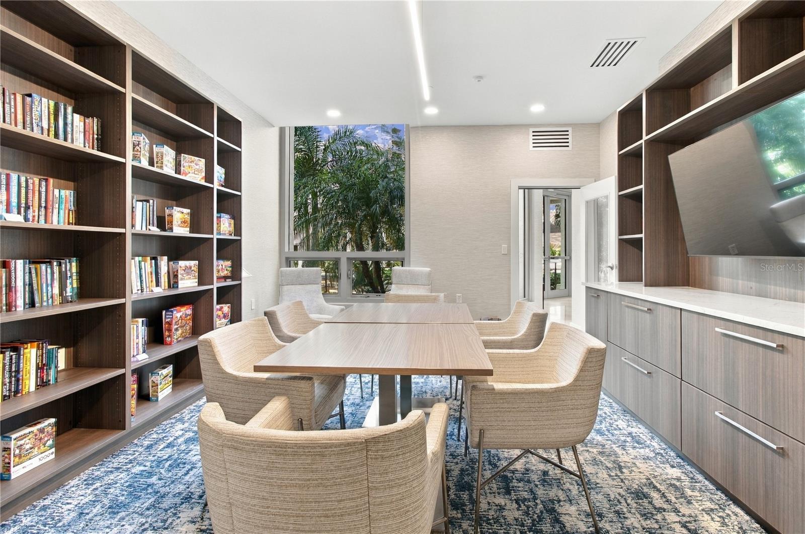 Furnished library meeting room with built-in bookshelves, a long table, and a tropical garden view.