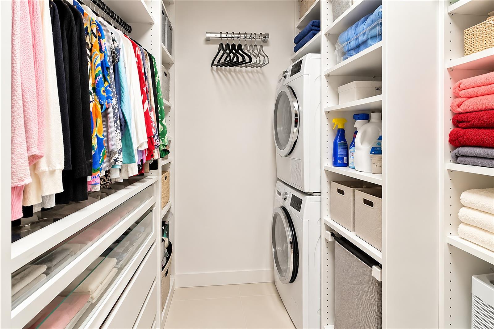 Walk-in laundry room with stacked washer and dryer, hanging clothes, folded towels, and built-in shelving.