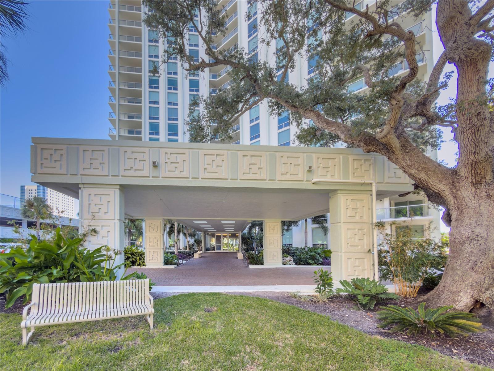 High-rise condominium entrance beneath a geometric canopy, framed by a mature oak and landscaped grounds.