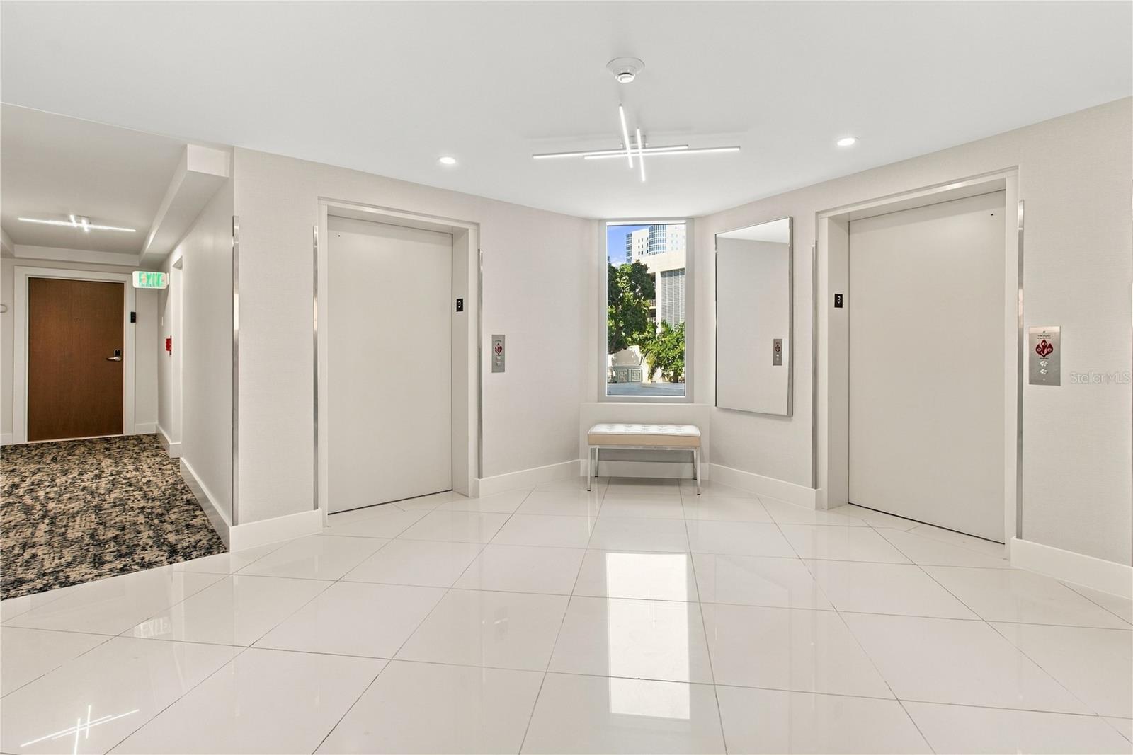 Condominium elevator lobby with two elevators, a window-side bench, and glossy white tile floors.