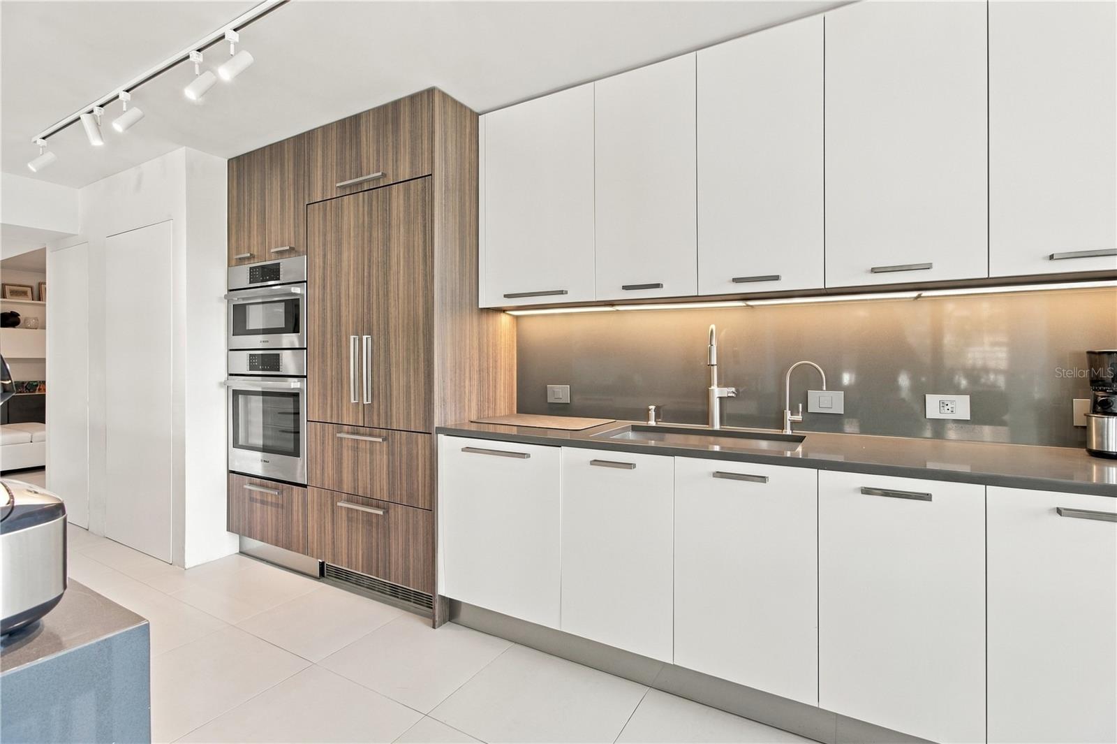 Modern kitchen with white cabinets, wood-grain cabinetry, built-in ovens, and warm under-cabinet lighting.
