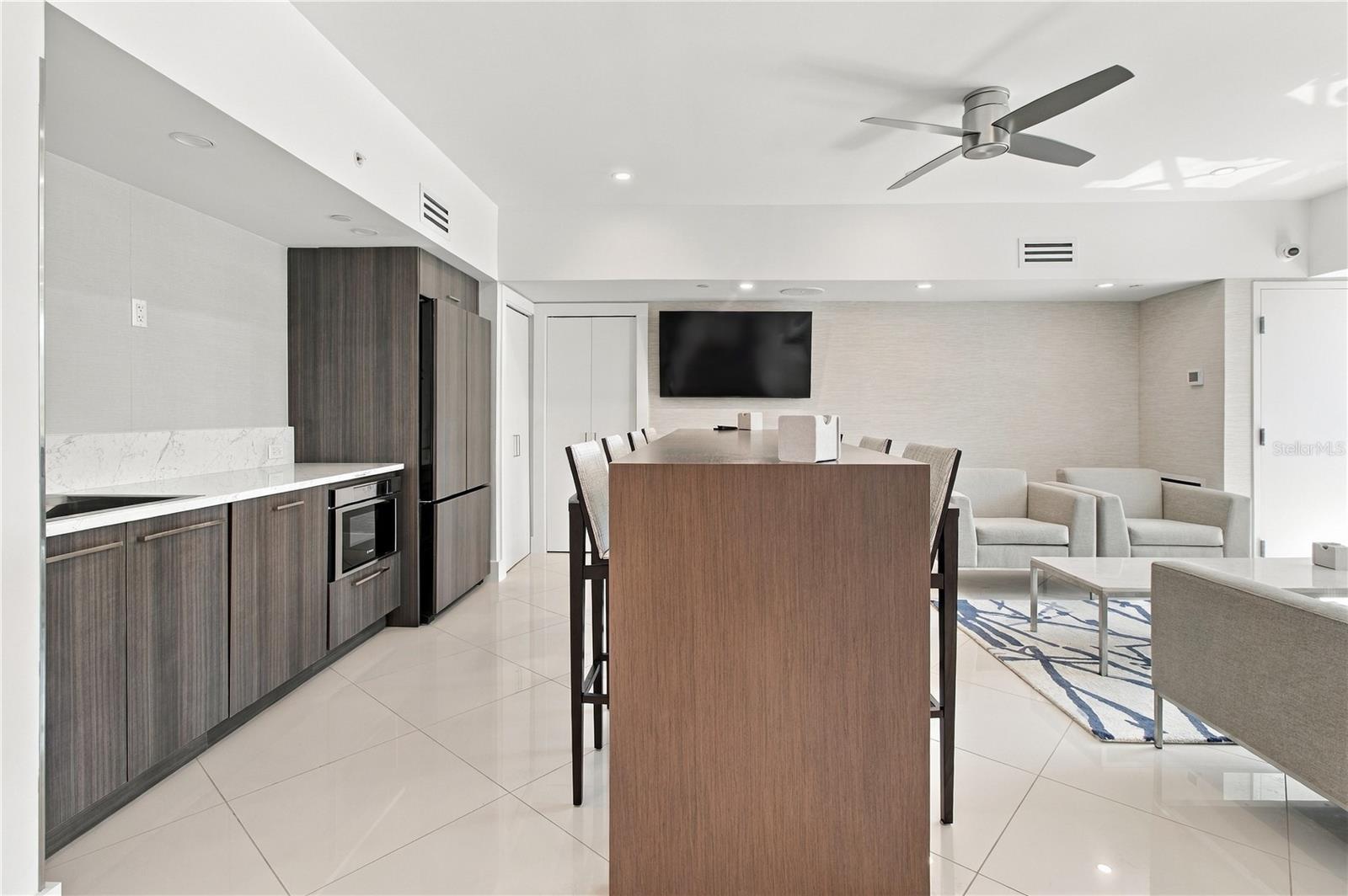 Open-plan condo kitchen and living area with dark wood cabinetry, a large dining island, and pale seating.