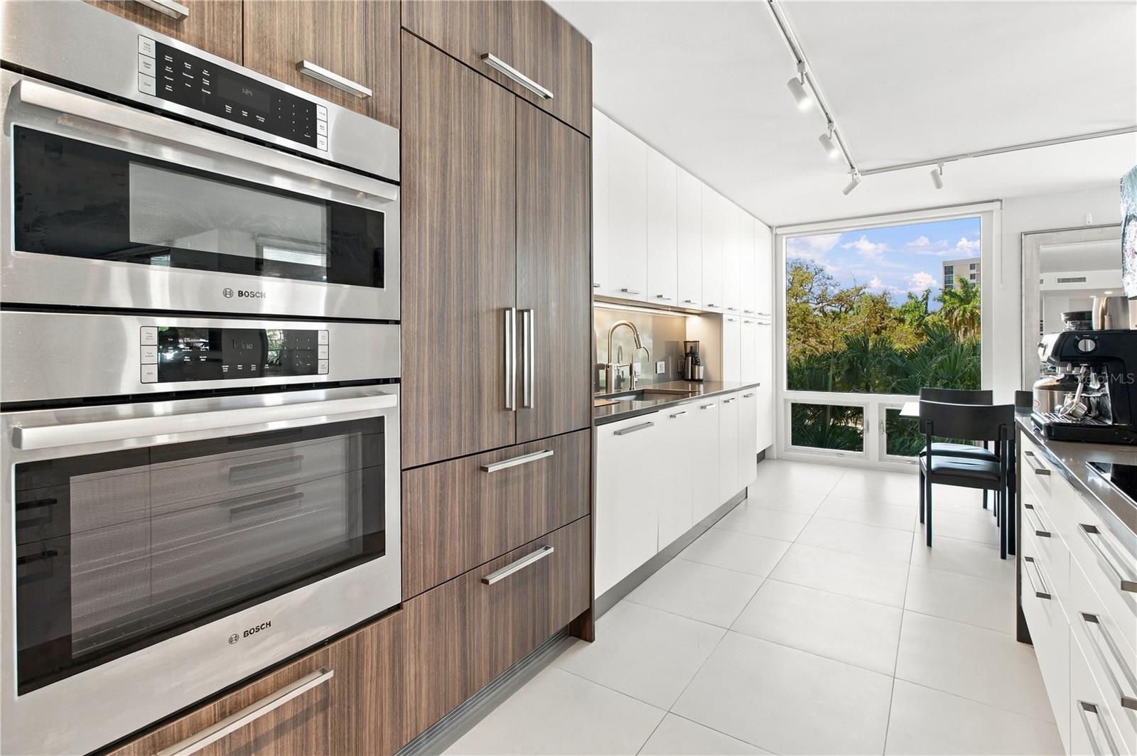 Modern kitchen with Bosch double ovens, wood-tone cabinetry, and a window overlooking palms.