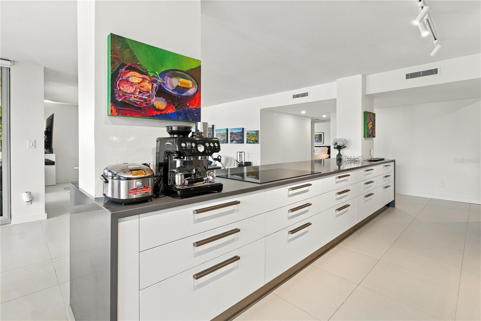 Modern white kitchen with a long dark-topped island, cooktop, and espresso machine.