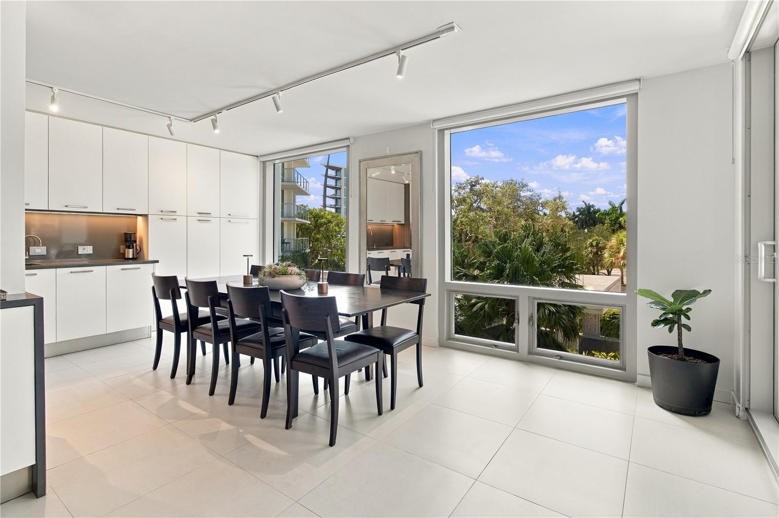 Modern condo dining area with white cabinetry, dark dining set, and large windows overlooking palm trees.