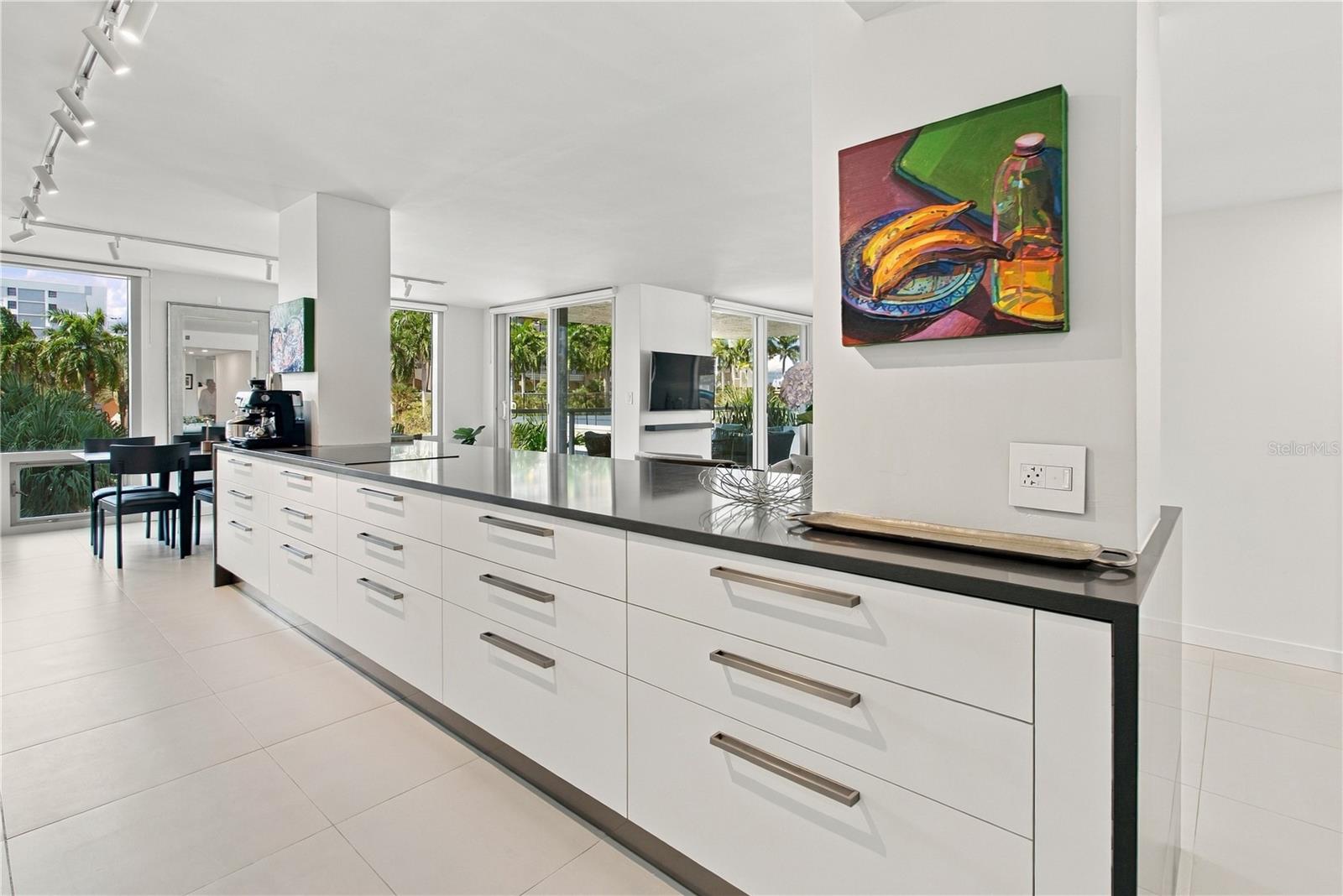 Modern white kitchen with a long island, dining nook, and windows overlooking tropical landscaping.