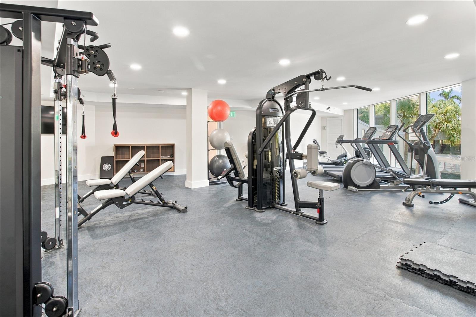 Fitness room with weight machines and cardio equipment beside large windows.