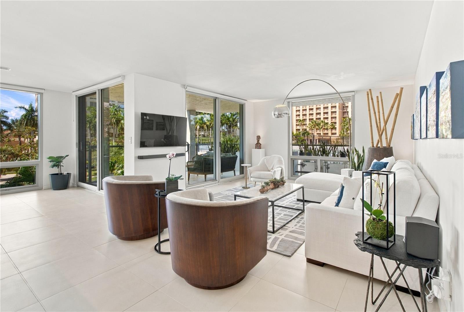 Staged living room with a white sectional, curved wood chairs, and large windows overlooking palm trees.