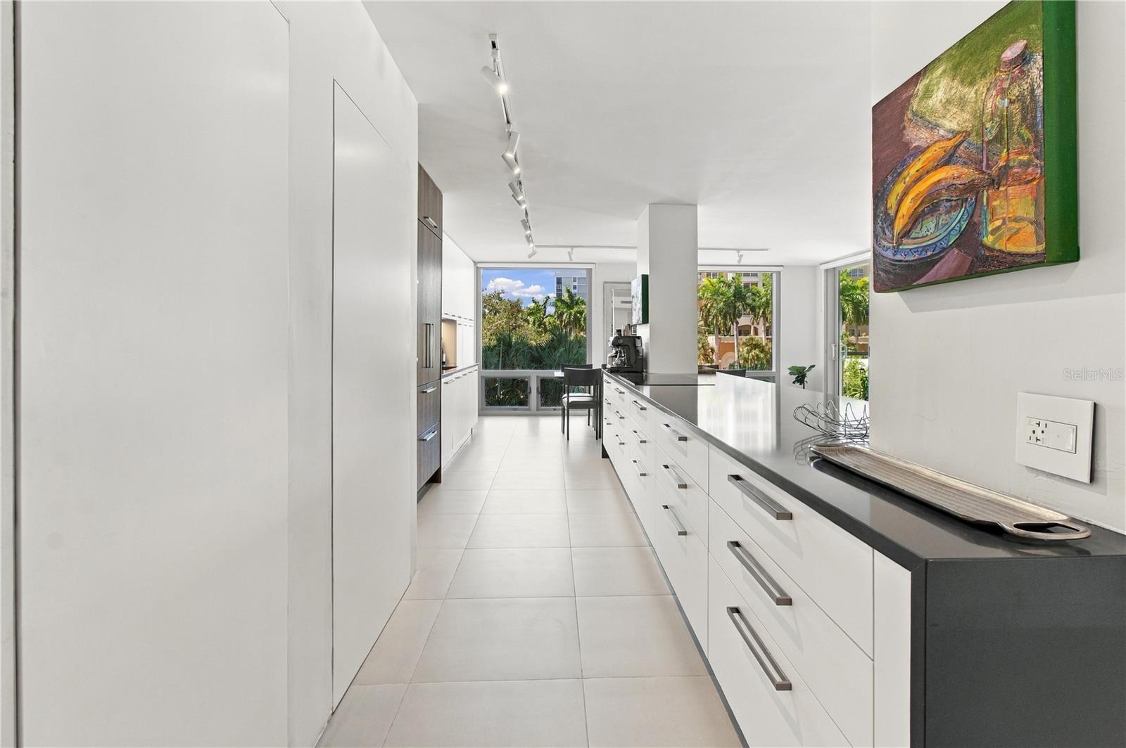 Modern white kitchen with a long dark countertop, large windows, and tropical greenery outside.