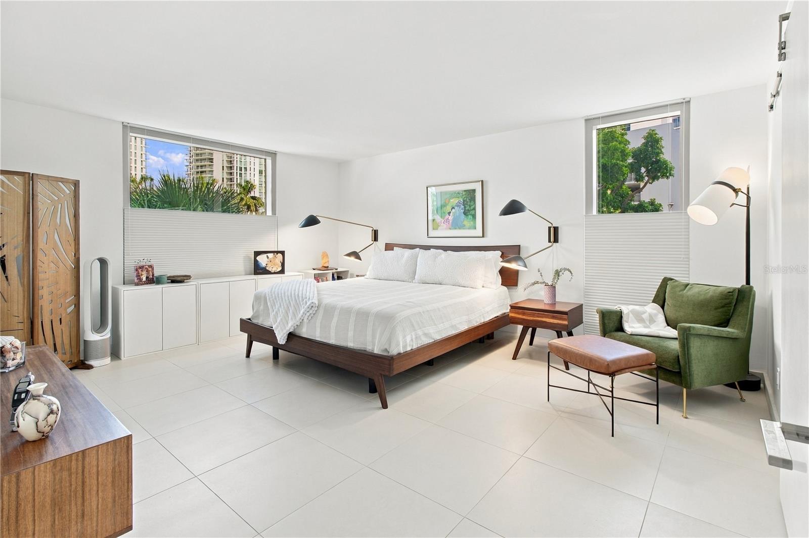 Bright bedroom with a large white bed, green armchair, and windows overlooking trees and nearby buildings.