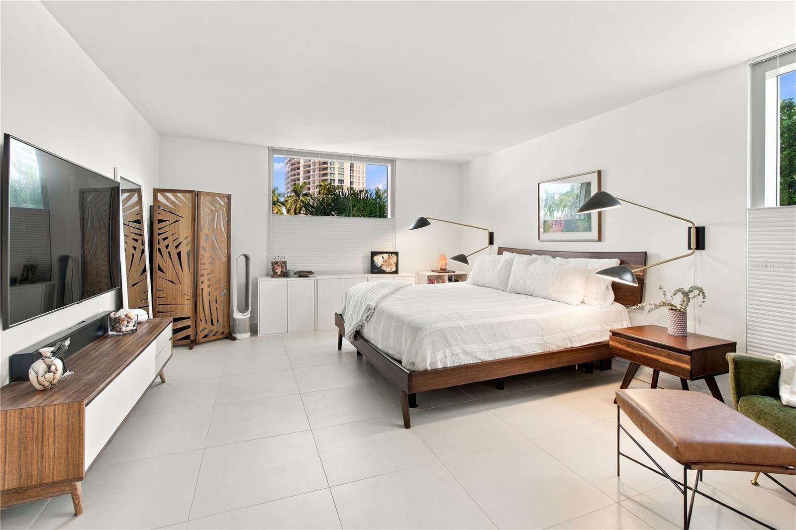 Furnished bedroom with a white bed, carved wooden screen, and windows overlooking palms and a high-rise.