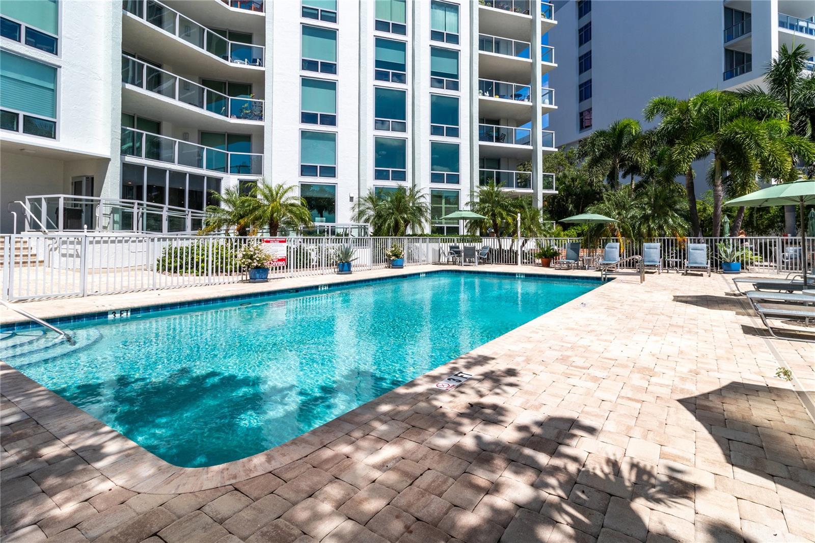 Condominium pool with a paved deck, palm trees, and lounge chairs beside a white high-rise.