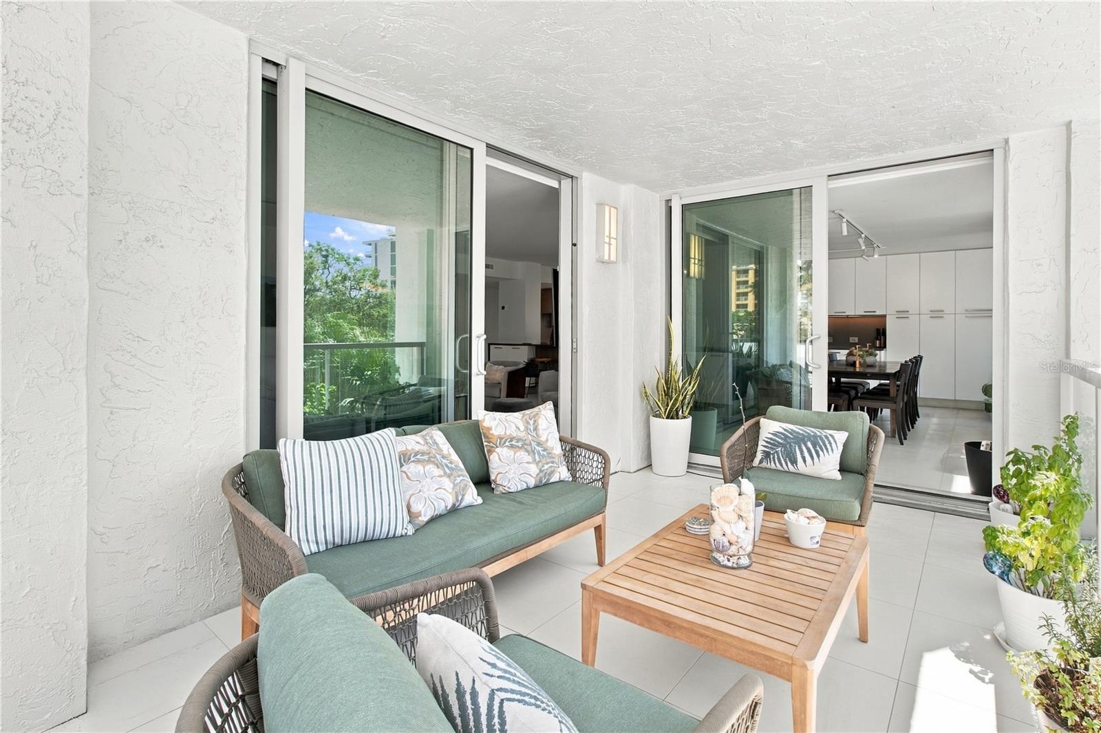 Covered condo balcony with green wicker seating, a wooden coffee table, and sliding doors to the dining area.