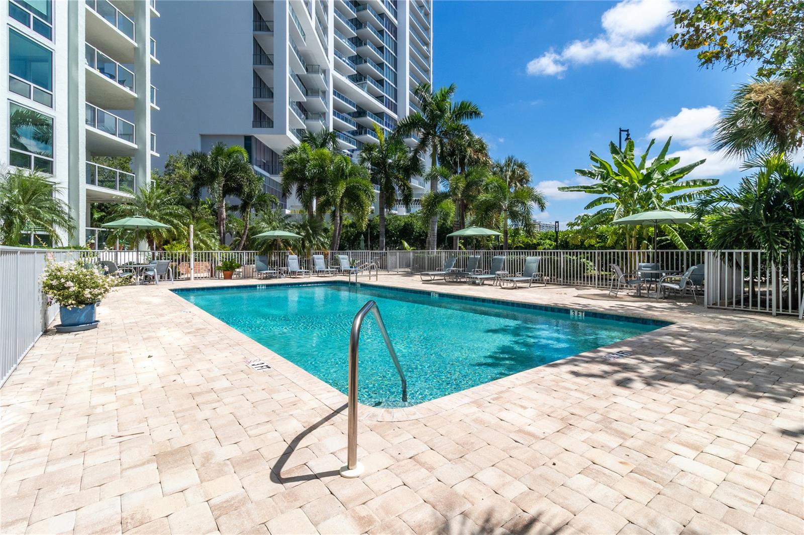 Condominium pool with a paved deck, lounge chairs, umbrellas, and palm trees.