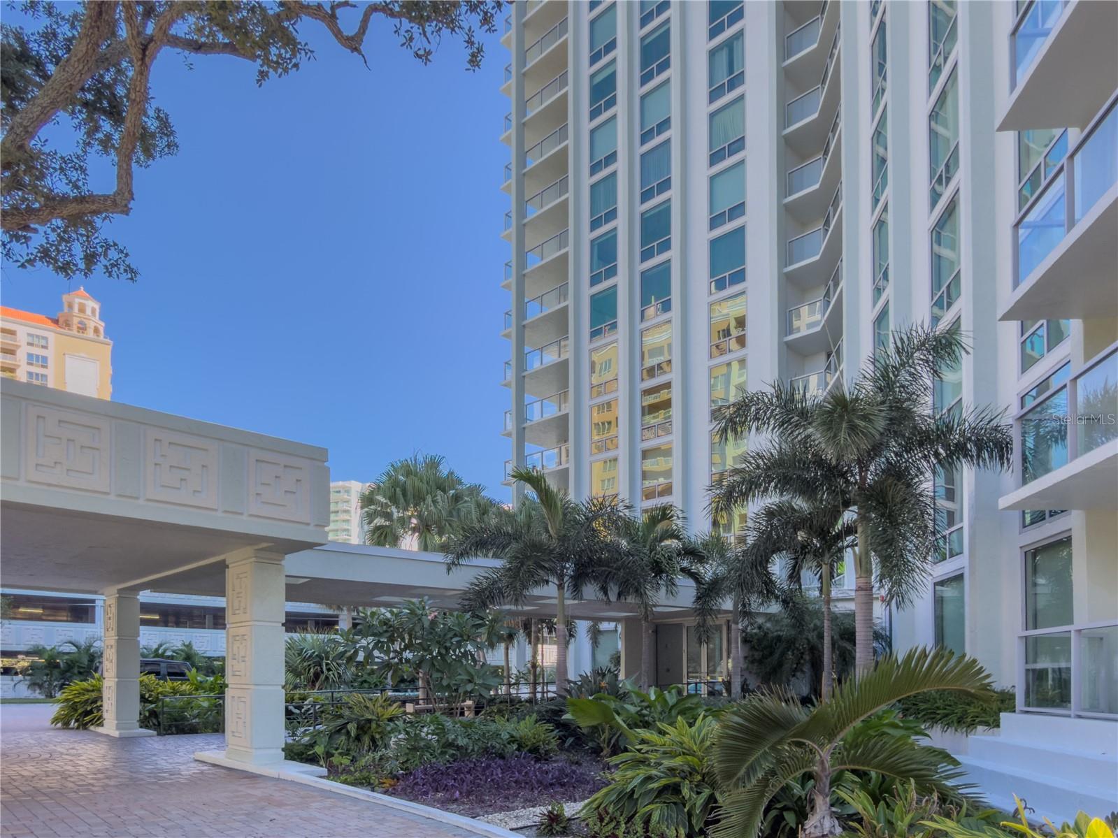 High-rise condominium exterior with balconies, a covered entrance, and tropical landscaping.