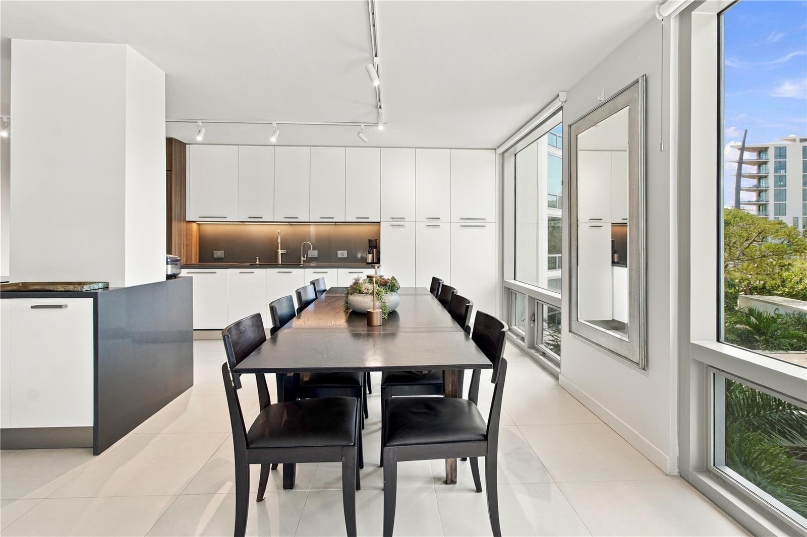 Modern condo kitchen and dining area with white cabinets, a long table, and floor-to-ceiling windows.
