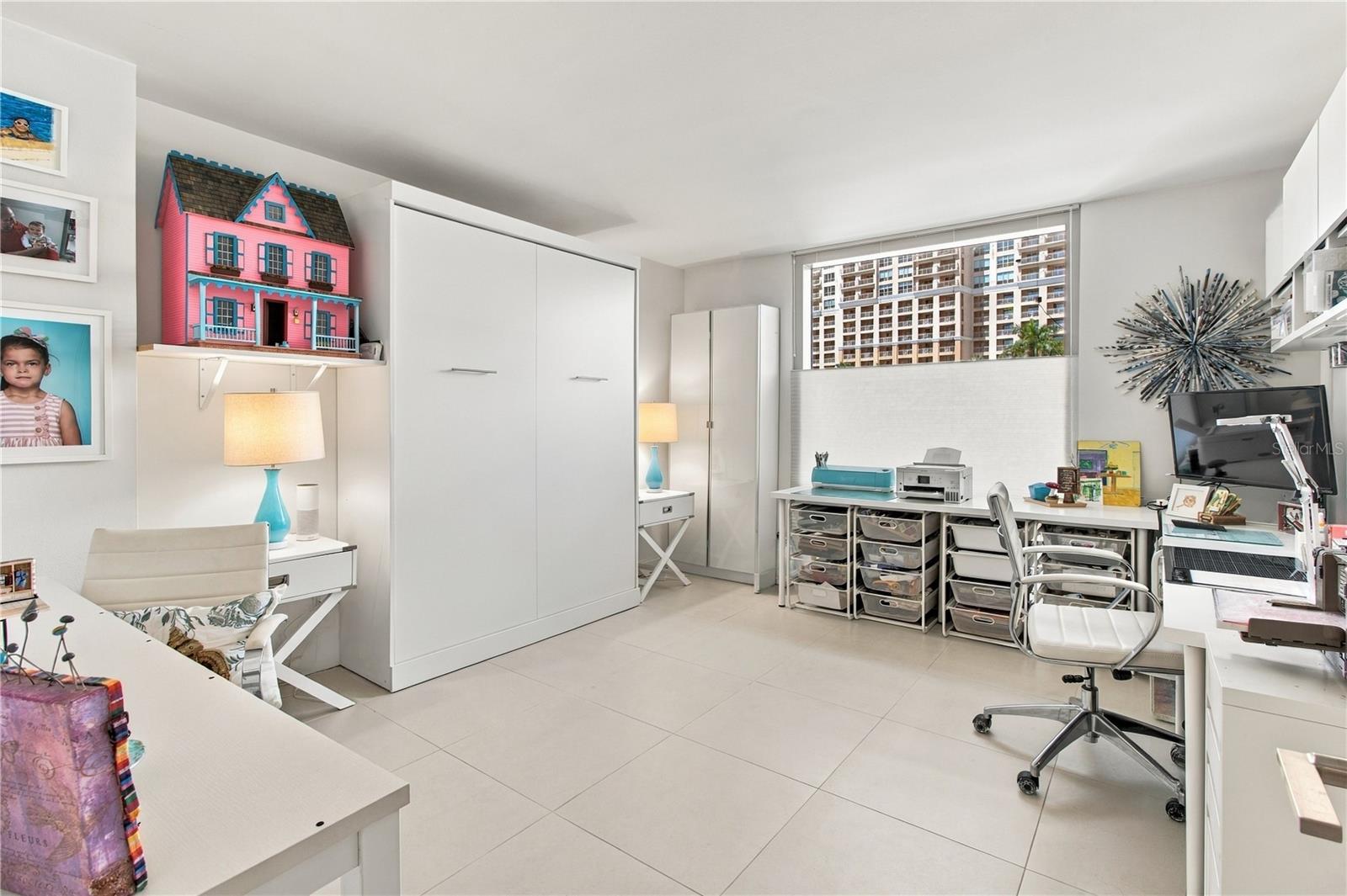 Condo office with a white wall bed, work desks, and a pink dollhouse beside a window overlooking high-rises.