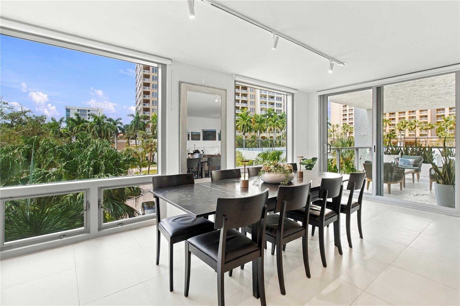 Dining area with a dark table and chairs, large windows, and views of palm trees and nearby buildings.