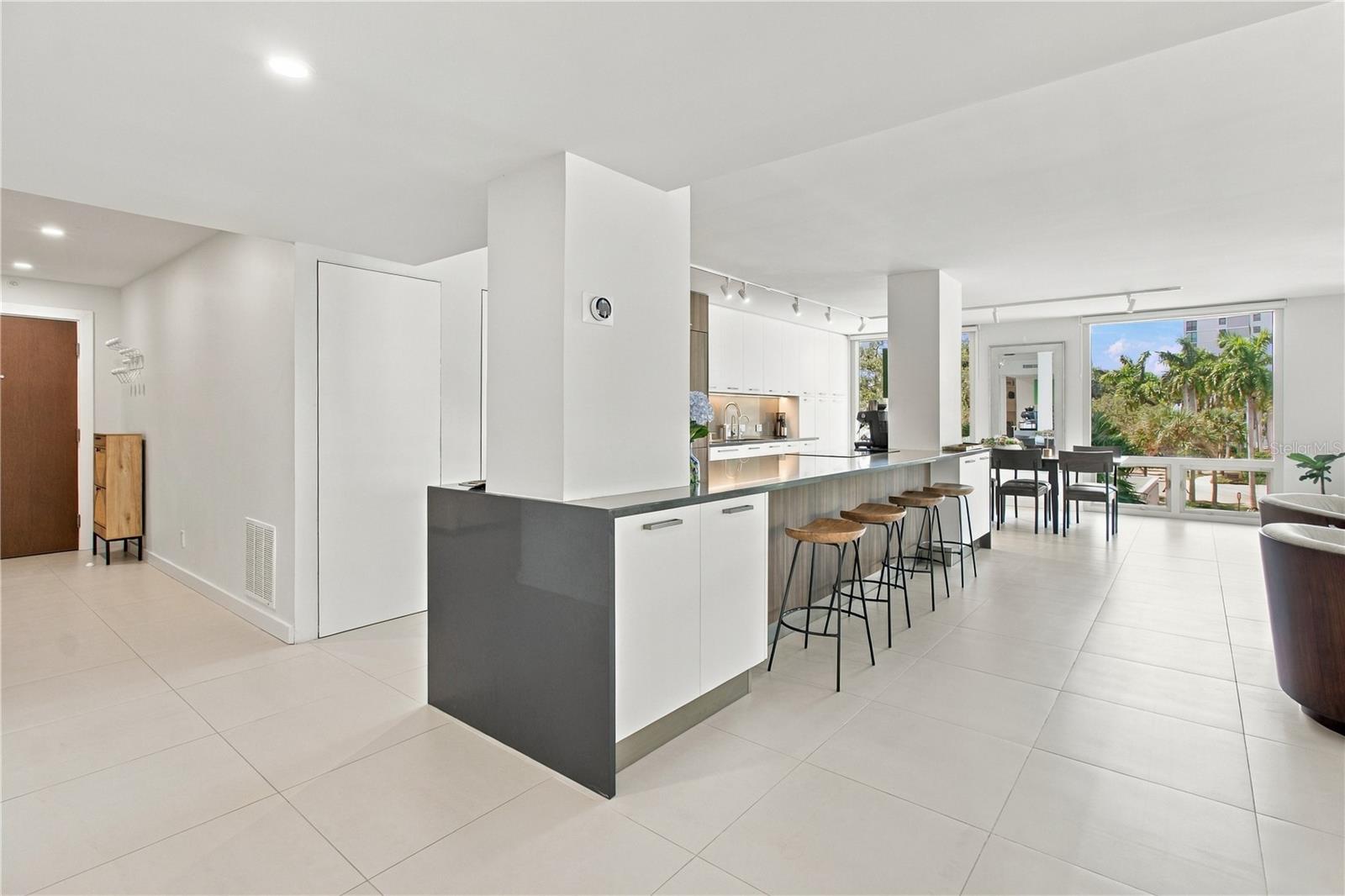 Open-plan condo kitchen with a long island and bar stools beside large windows.
