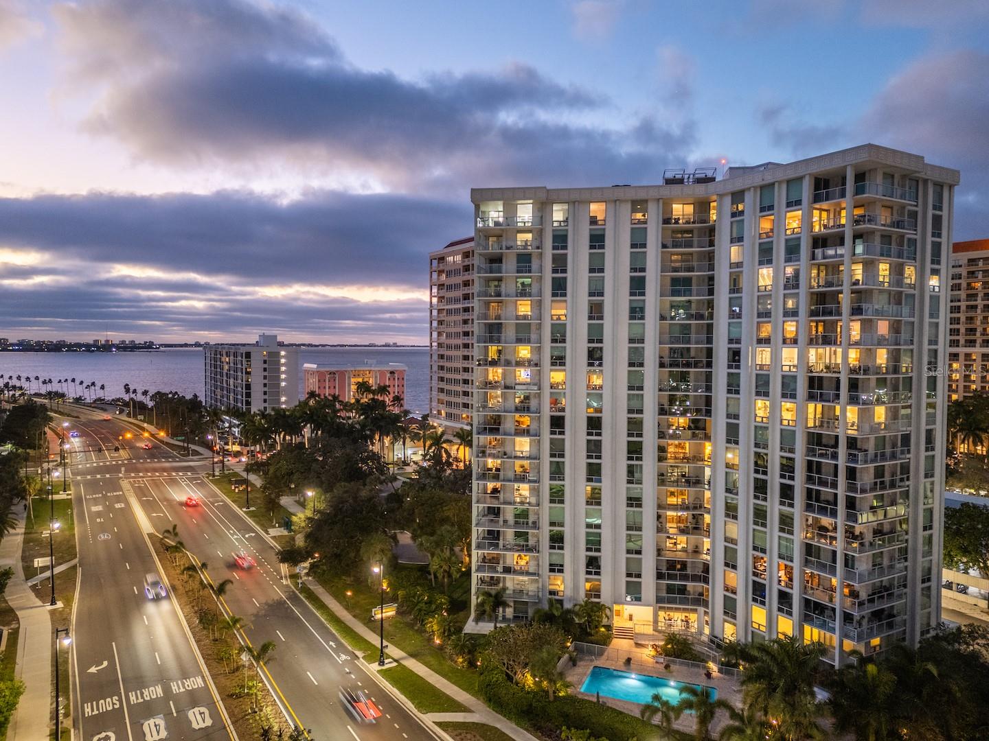Condominium tower at dusk beside the bay, with balconies, a palm-lined road, and a swimming pool.