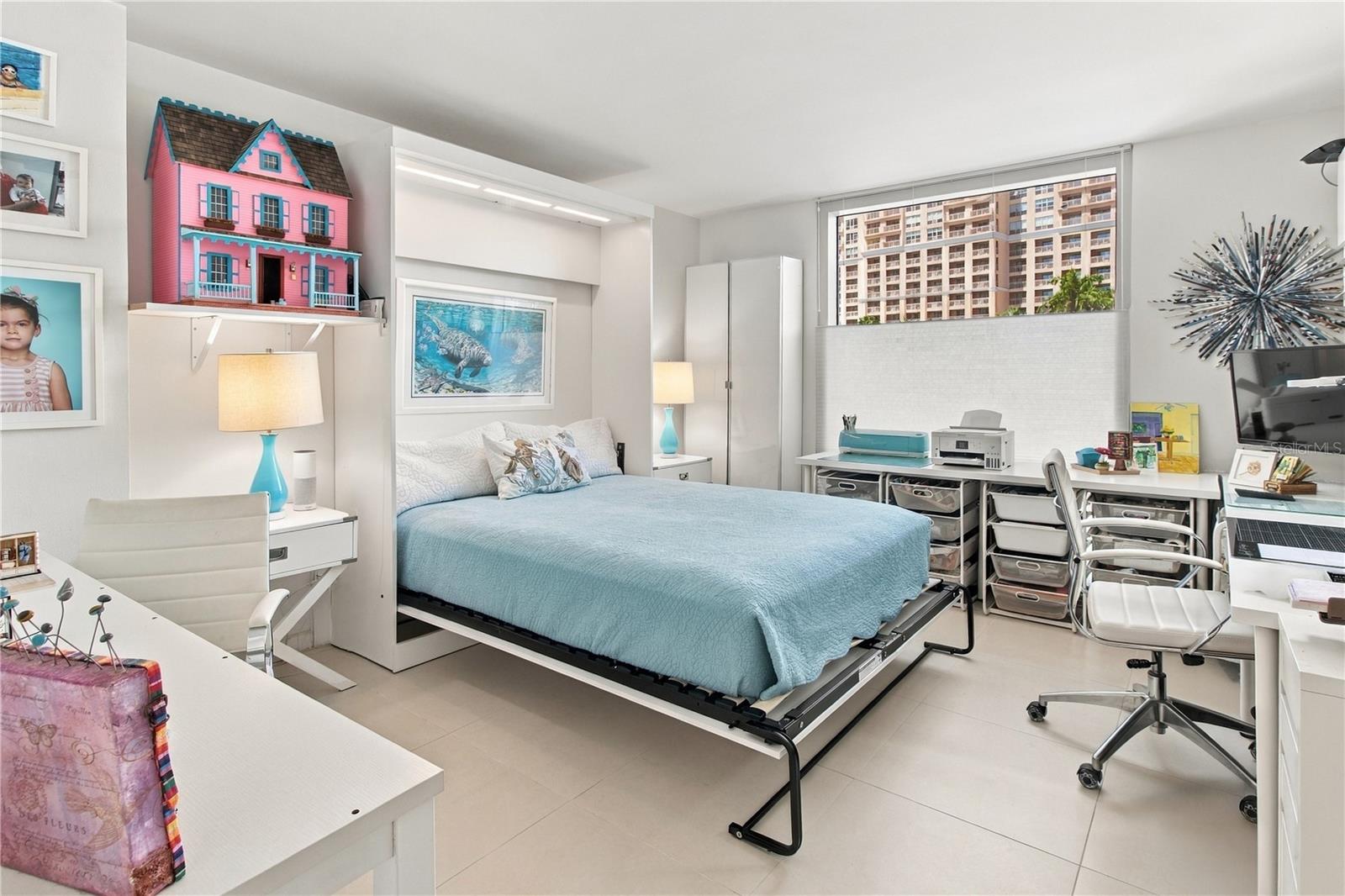 Bright bedroom with an aqua bed, white workspaces, a pink dollhouse, and a high-rise view.