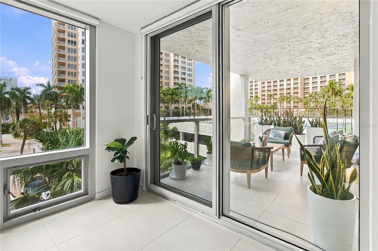 Condo interior opening onto a furnished balcony with potted plants, palm trees, and neighboring high-rises.