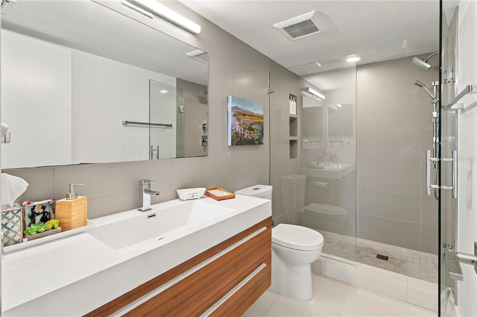 Modern bathroom with a wood-front vanity, large mirror, and glass-enclosed gray-tile shower.