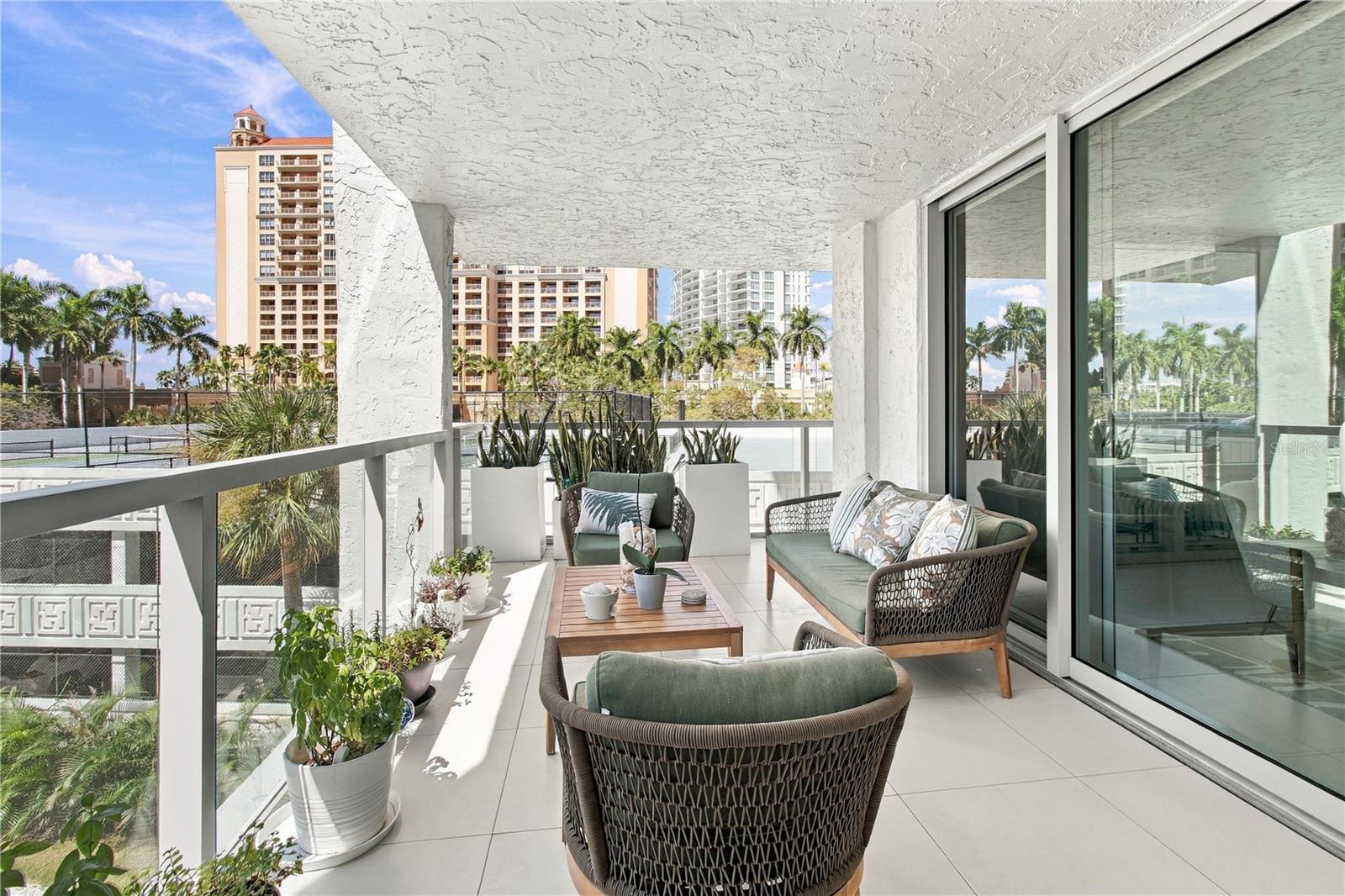 Furnished condo balcony with outdoor seating, potted plants, palm trees, and neighboring high-rises.