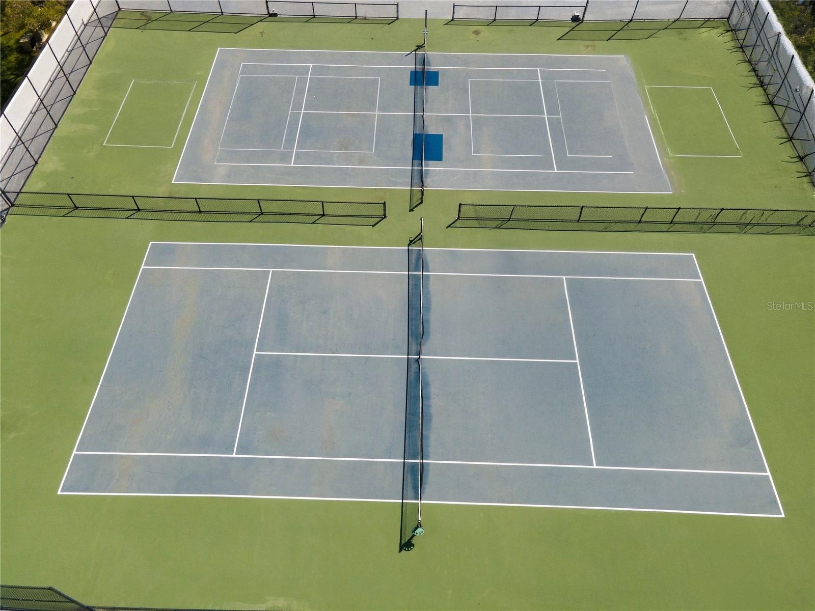 Two fenced outdoor tennis courts viewed from above, with blue-gray surfaces and green surrounds.